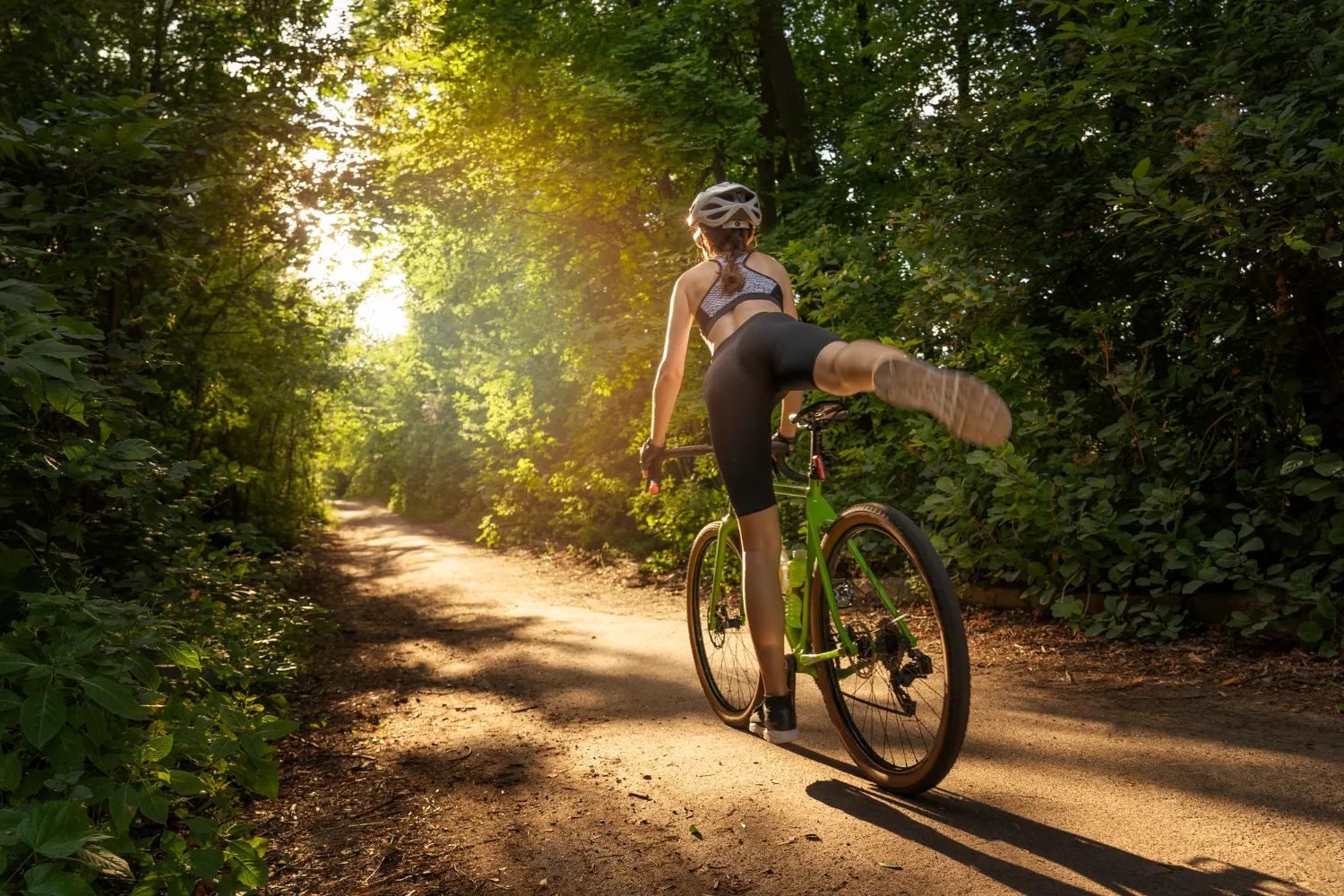Ciclista femenina realizando una acrobacia sobre una bicicleta verde en un sendero rodeado de árboles, con luz solar filtrándose a través de la vegetación.